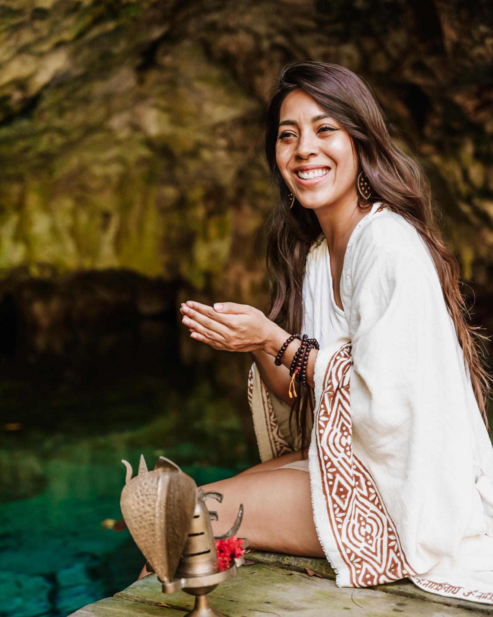 La Dra. Pamela Corona sonríe sentada a la orilla de un cenote, con las manos juntas, junto a un lingam de Shiva de latón