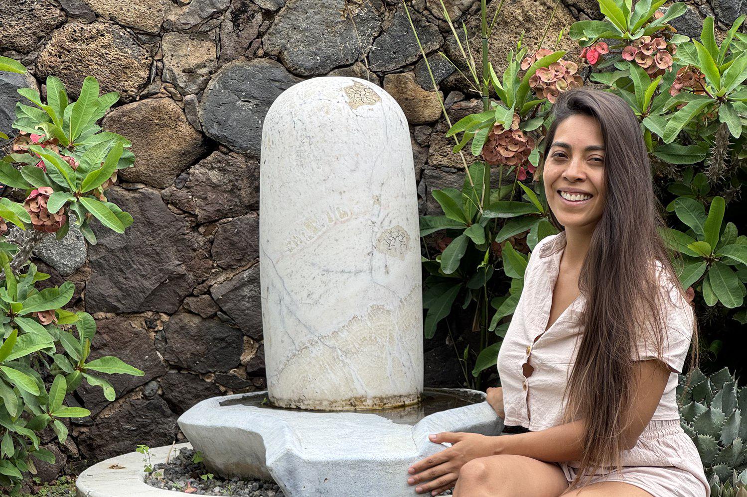 La Dra. Pamela Corona sentada junto a un lingam de piedra en un jardín