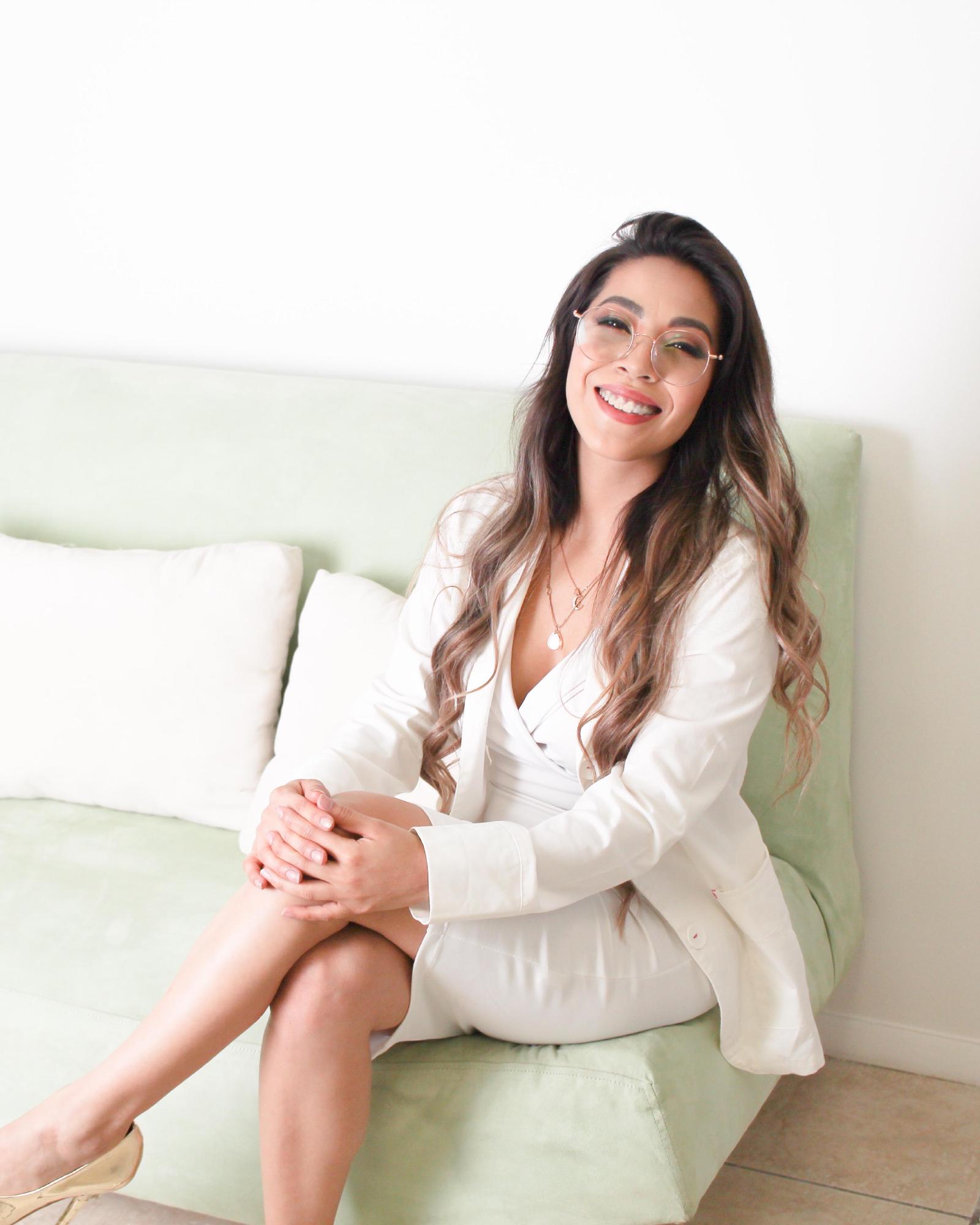 La Dra. Pamela Corona, con traje blanco y lentes, sonríe sentada en un sillón verde claro