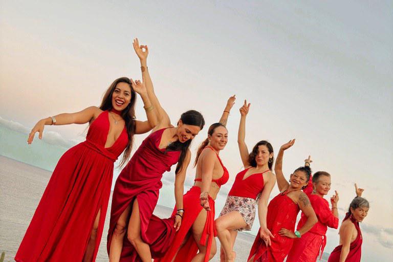 Un grupo de mujeres danzando con vestidos rojos a la orilla del agua al atardecer